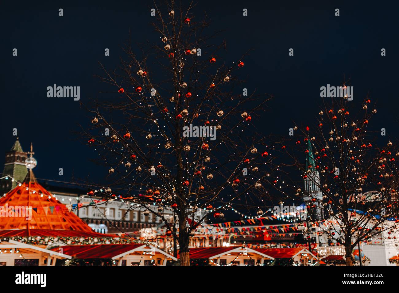 Tree branches decorated with red and gold shiny Christmas balls. New Year outdoor decorations against a blue evening sky and glowing building. Christm Stock Photo