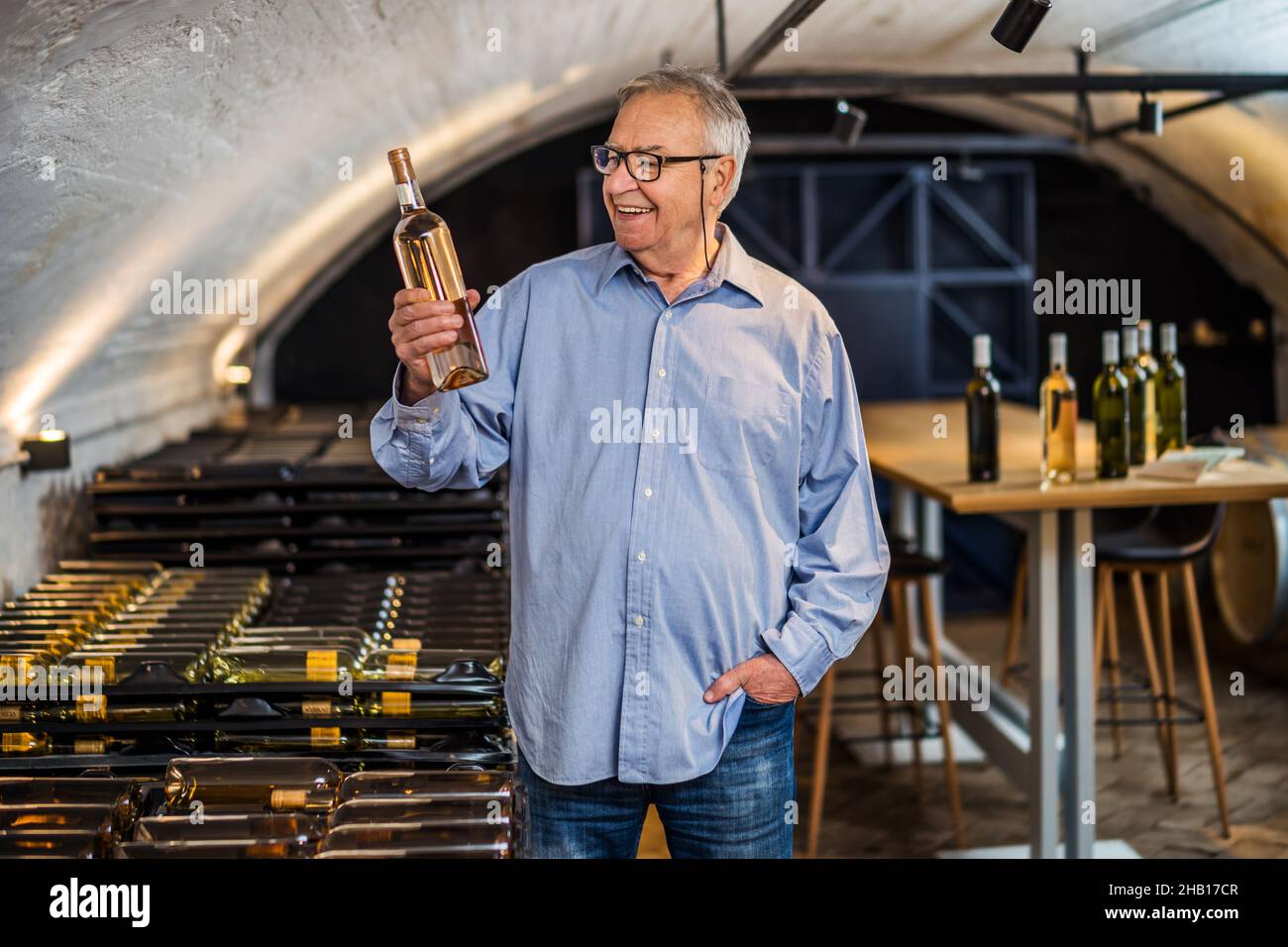 Portrait of happy senior man who owns winery. He is holding bottle of wine in his wine cellar