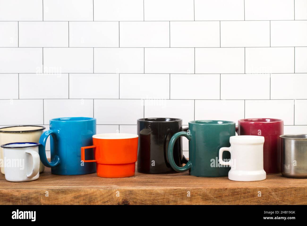 Multicolored mugs on a wooden kitchen board in front of a subway tiles wall Stock Photo