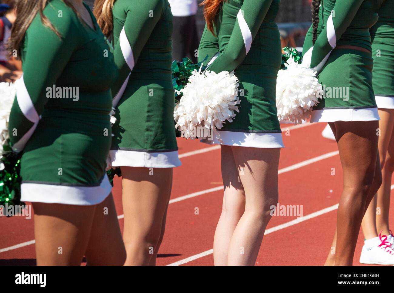 Close up of high school cheerleaders standing on a track watching the football game holding their pom poms behind their backs. Stock Photo
