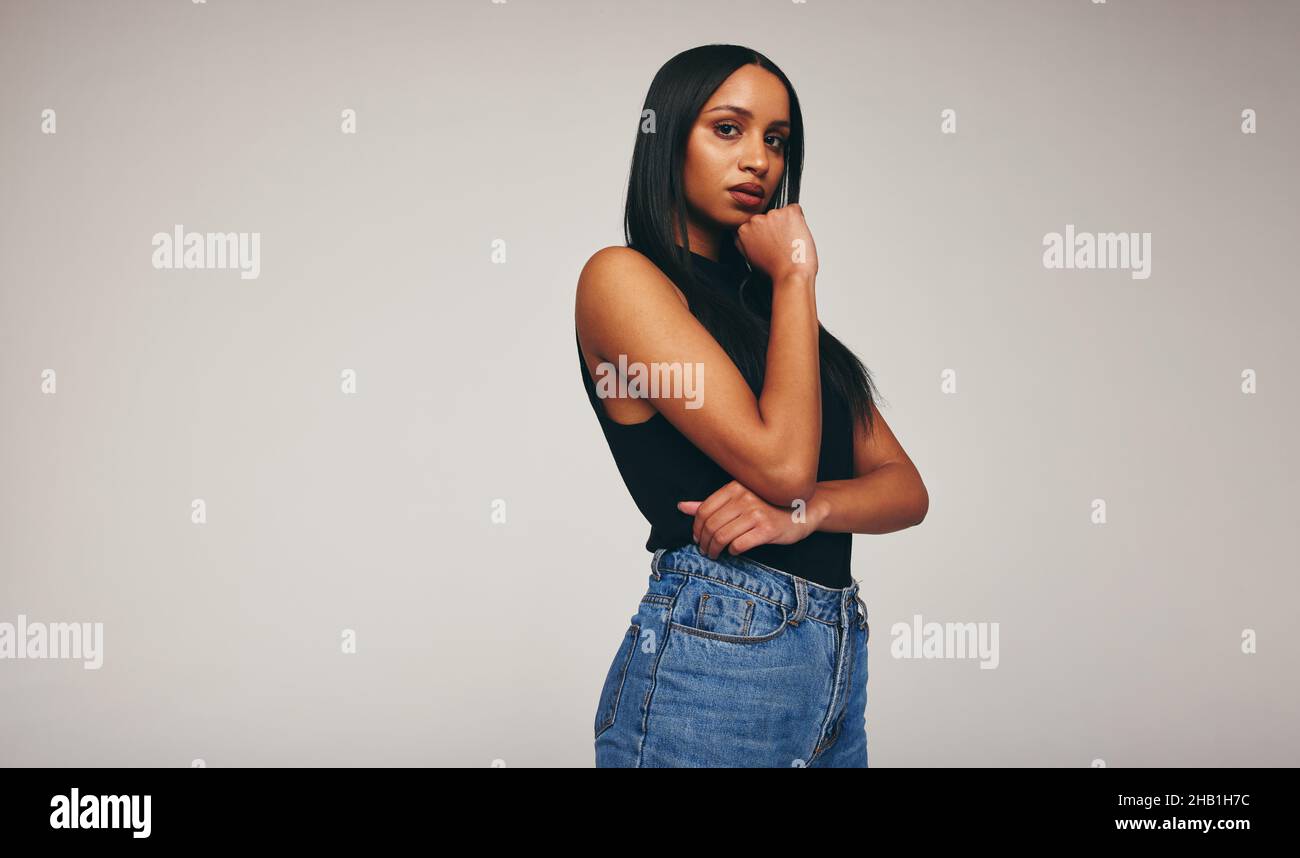 Self-confident young woman looking at the camera while standing alone against a grey background. Fashionable young woman wearing denim jeans in a stud Stock Photo