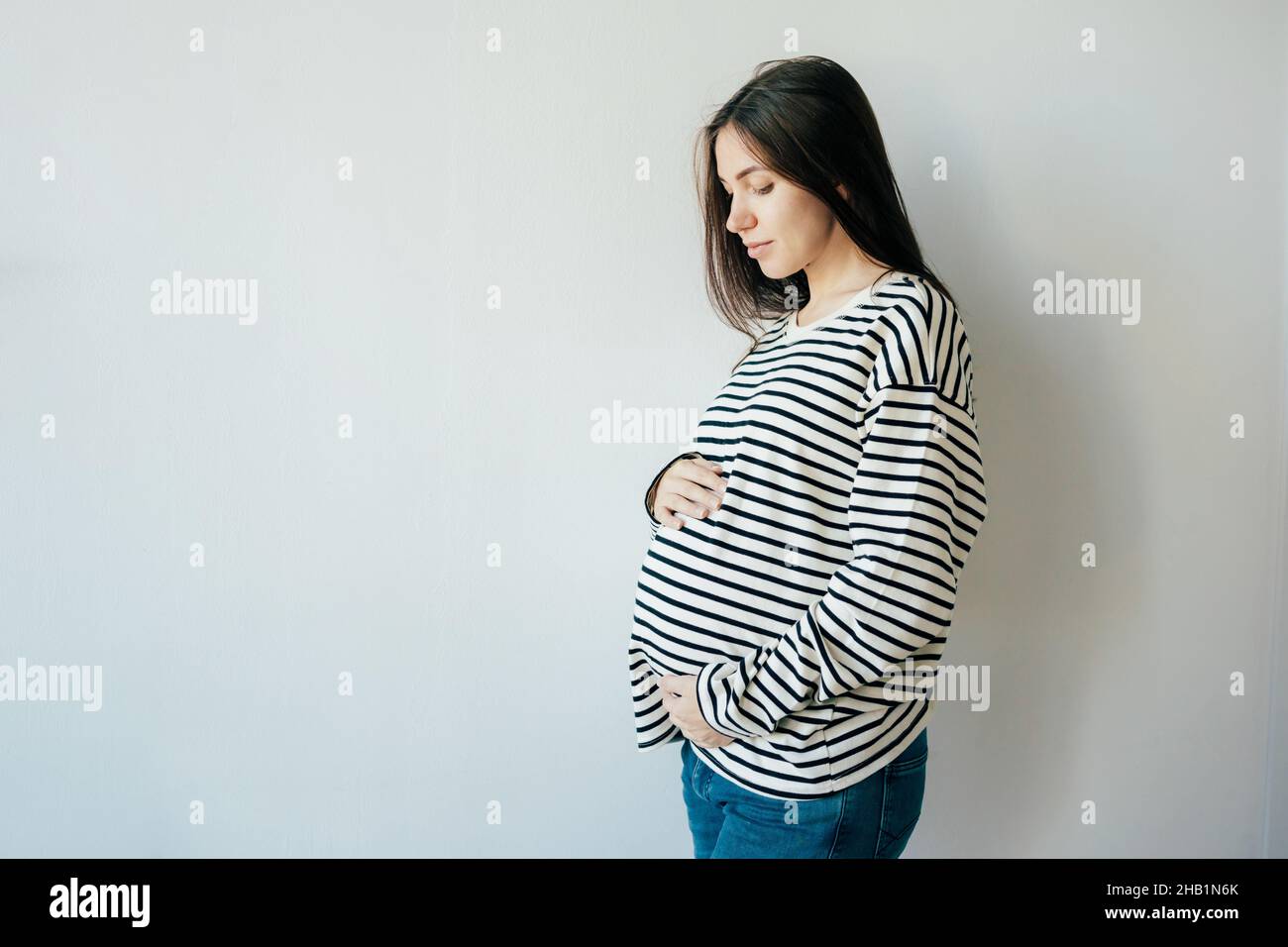 Tender young pregnant woman in a striped sweatshirt against the background of a white wall. Stock Photo
