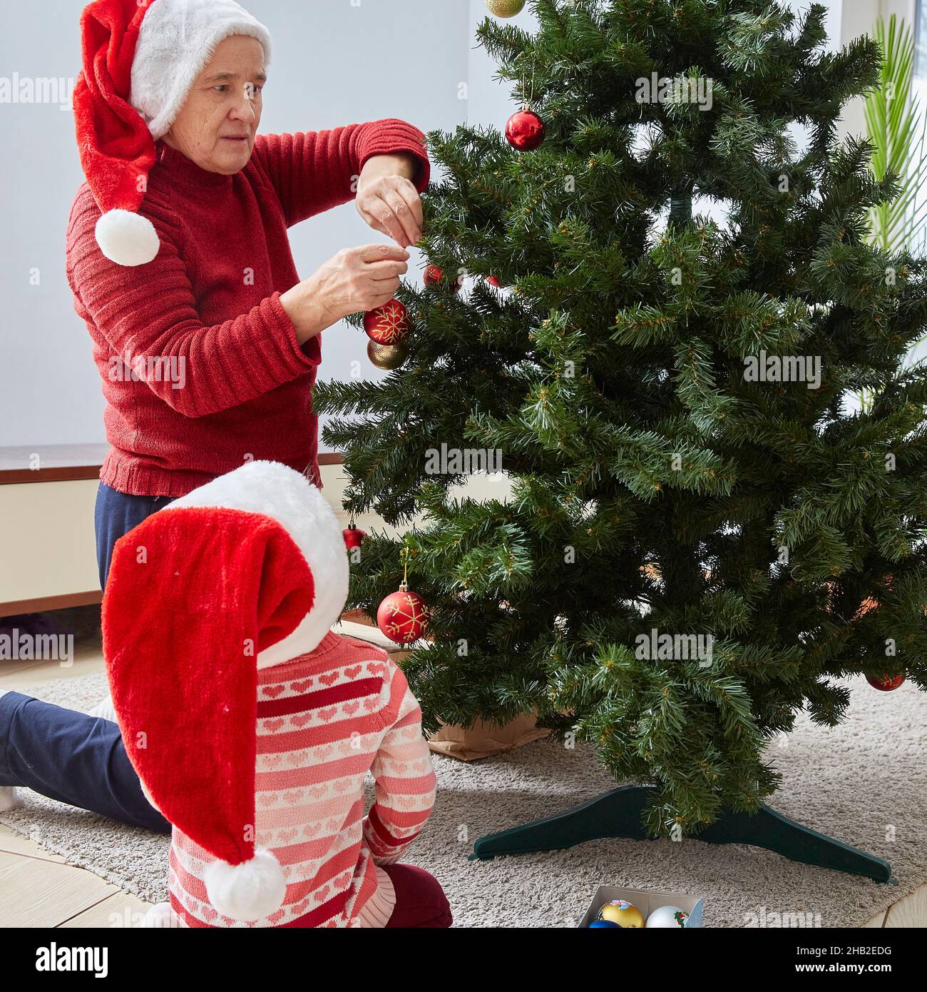 Grandma is decorating the Christmas tree for the New year and Christmas. The child looks at the grandmother Stock Photo