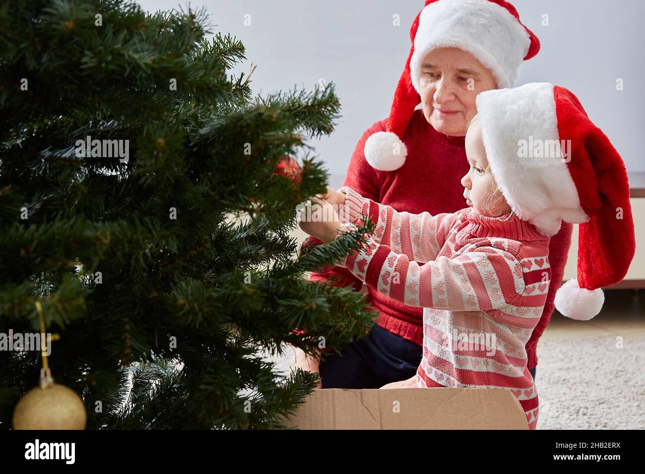 little girl hangs decorations on the Christmas tree. Grandmother admires her granddaughter. The concept of family and New Year's Christmas holidays Stock Photo