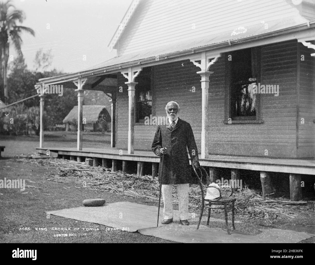 King George of Tonga (aged 86), Burton Brothers studio, photography studio, 26 July 1884, New Zealand, black-and-white photography, A man with grey hair and beard, formally dressed in dark suit coat and white trousers, with walking stick in right hand and left hand on a chair, is standing on a weaved mat in front of a colonial building. A Fale is in the background Stock Photo