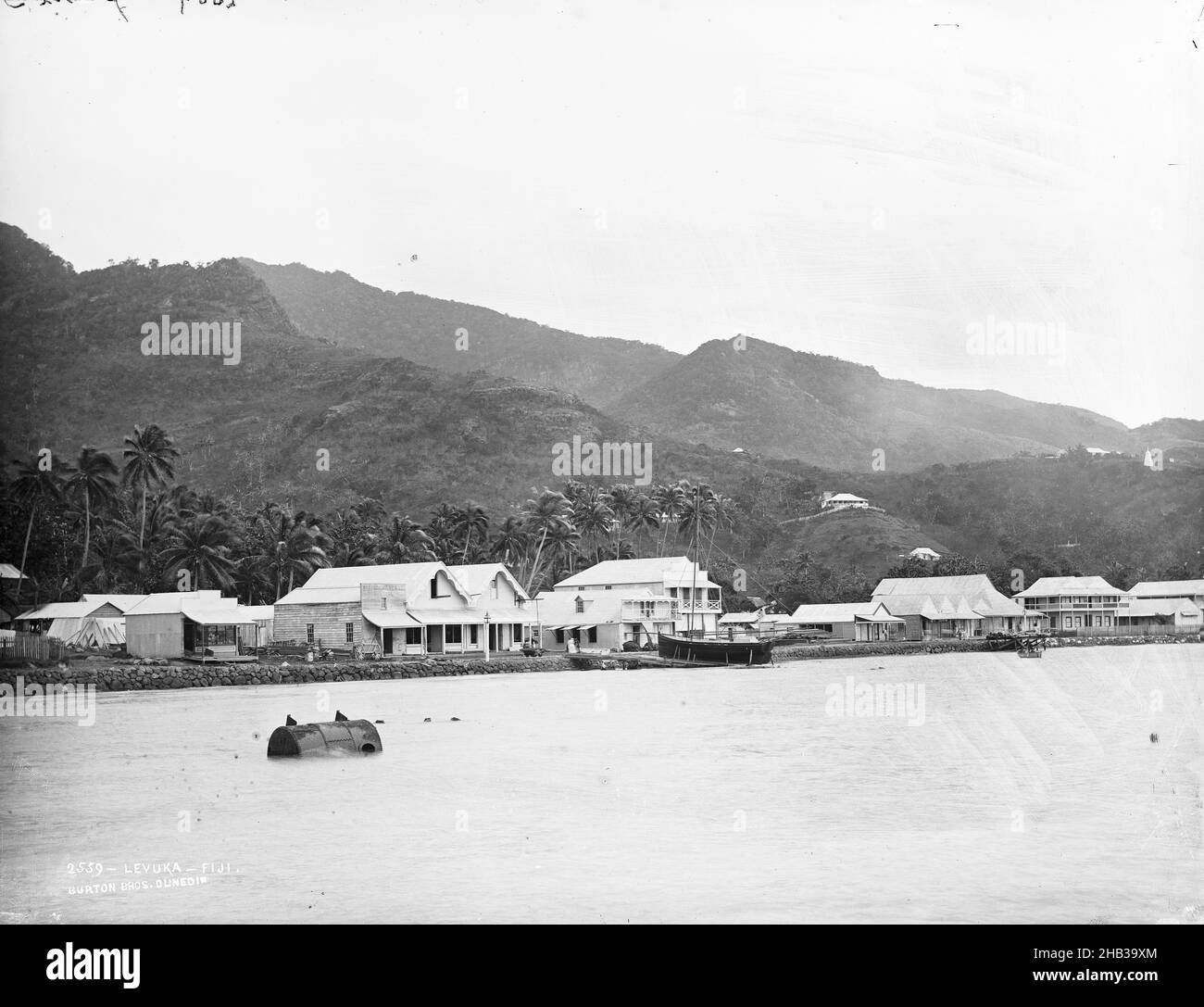 Levuka, Fiji, Burton Brothers studio, photography studio, 14 July 1884, New Zealand, gelatin dry plate process, Second plate in three plate panorama taken from the jetty. Foreground sea running to seawall with road above, buildings on roadside, coconut trees behind buildings (from centre to left) and steep hill rising behind buildings. To right a cylindrical buoy floats in the sea, a longboat is sitting on the shoreline. The hill line drops away left to right Stock Photo