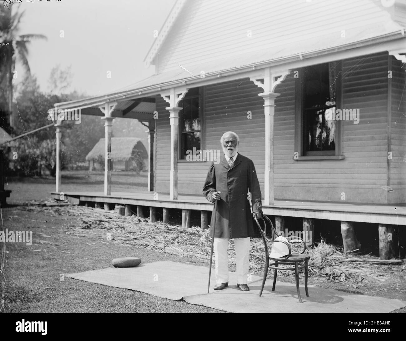 [King George of Tonga], Burton Brothers studio, photography studio, 26 July 1884, New Zealand, gelatin dry plate process, A man with grey hair and bed, formally dressed in dark suit coat and white trousers, with walking stick in right hand and left hand on a chair, is standing on a weaved mat in front of a colonial building. A Fale is in the background Stock Photo