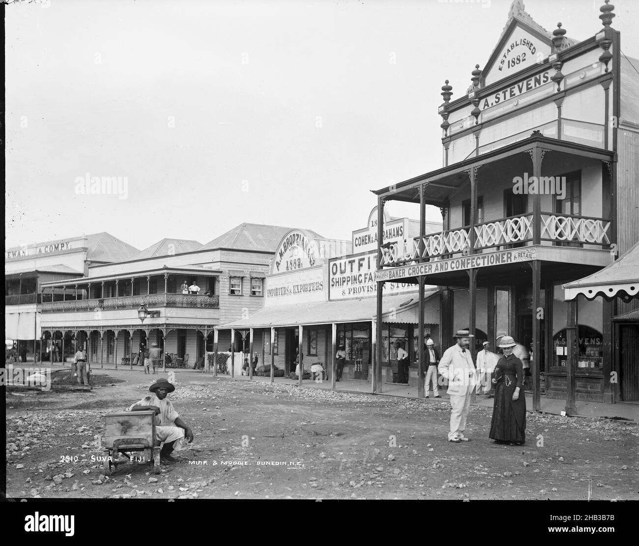 Suva, Fiji, Burton Brothers studio, photography studio, 1884, New Zealand, gelatin dry plate process, A populated Suva street lined with businesses. Two of these were established in 1882, just two years before Burton took this photograph. The shops reflect the needs of a growing market of European tourists. Shopfronts include 'Cohen & Abrahams Outfitters' - a shipping supplies and provisions store, 'A. M. Brodziak & Co. Importers and Exporters', and 'A. Stevens - Baker, Grocer & Confectioner Stock Photo