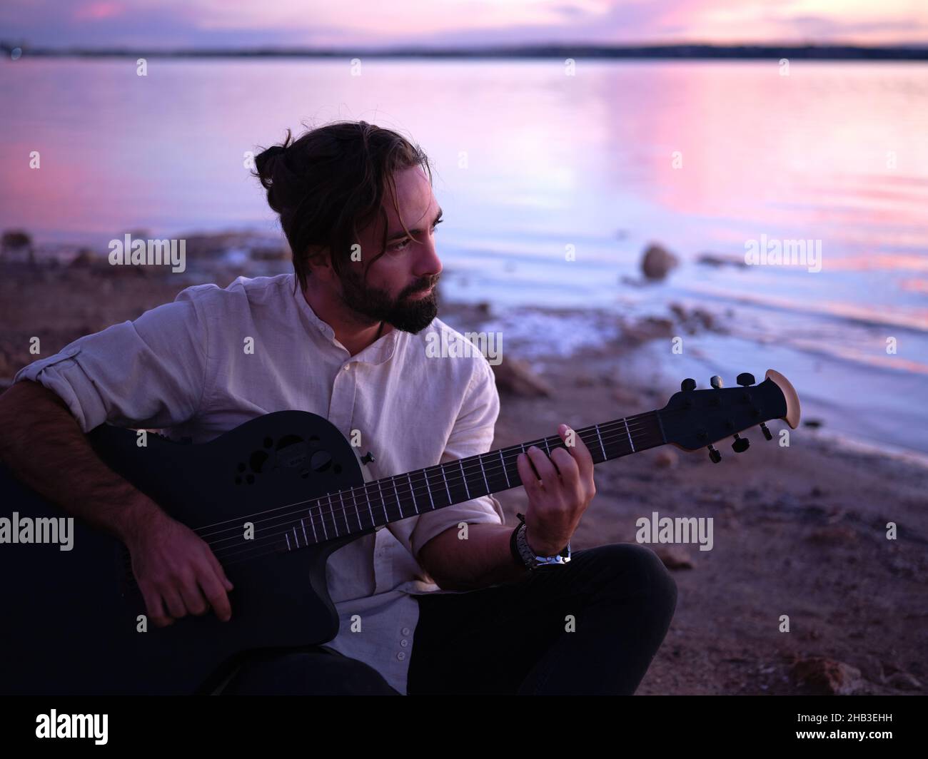 guitarist sitting by a lake reflecting the pink sunset light Stock Photo