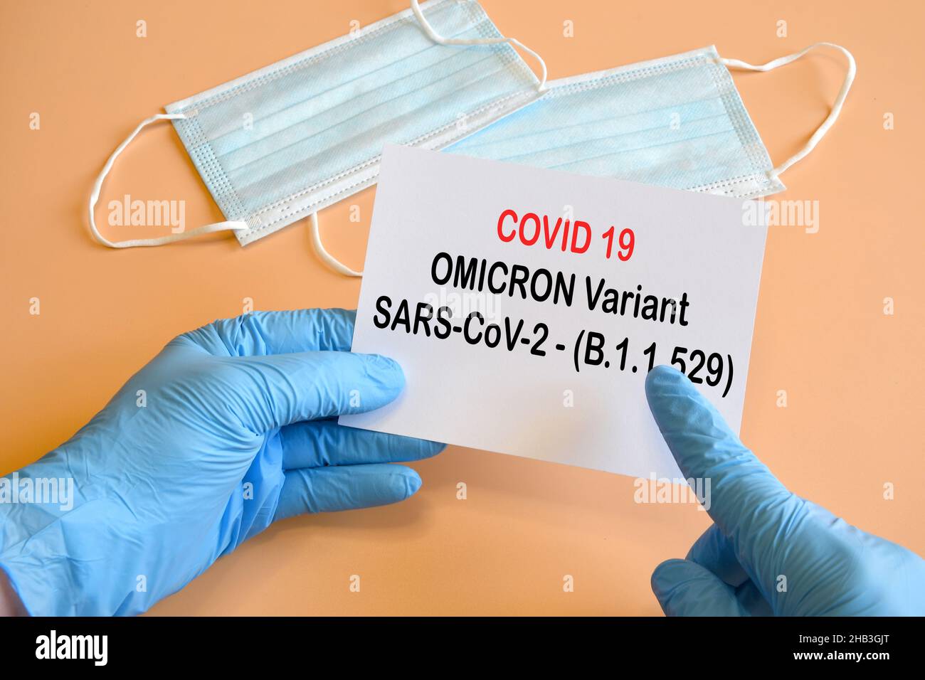The hand of the doctor in blue gloves holds a white paper with text 'COVID 19 Omicron variant' with medical protection masks on background. Stock Photo