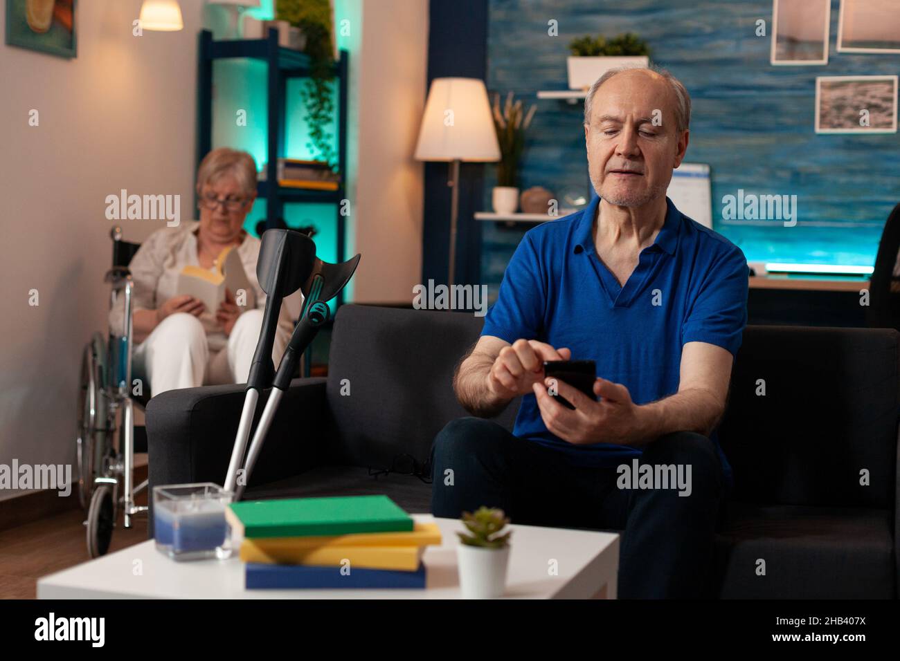 Aged caucasian man using smartphone on living room sofa. Senior adult holding modern device with technology looking at screen while old woman in wheelchair sitting in background Stock Photo
