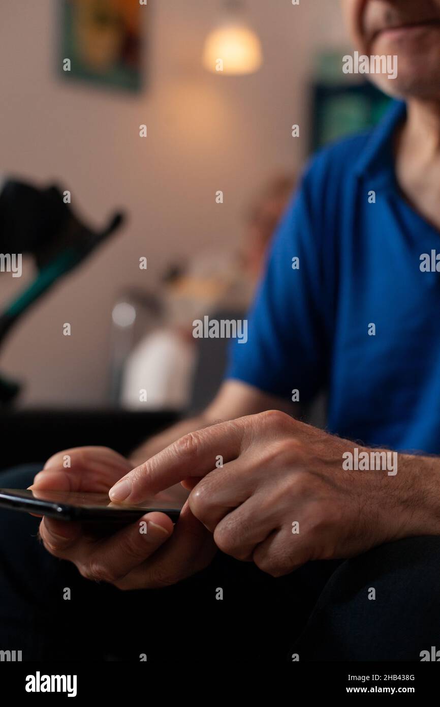 Close up of unrecognisable aged hands holding smartphone at home on sofa. Senior retired man touching device with modern technology while sitting on couch in living room. Person using gadget. Stock Photo