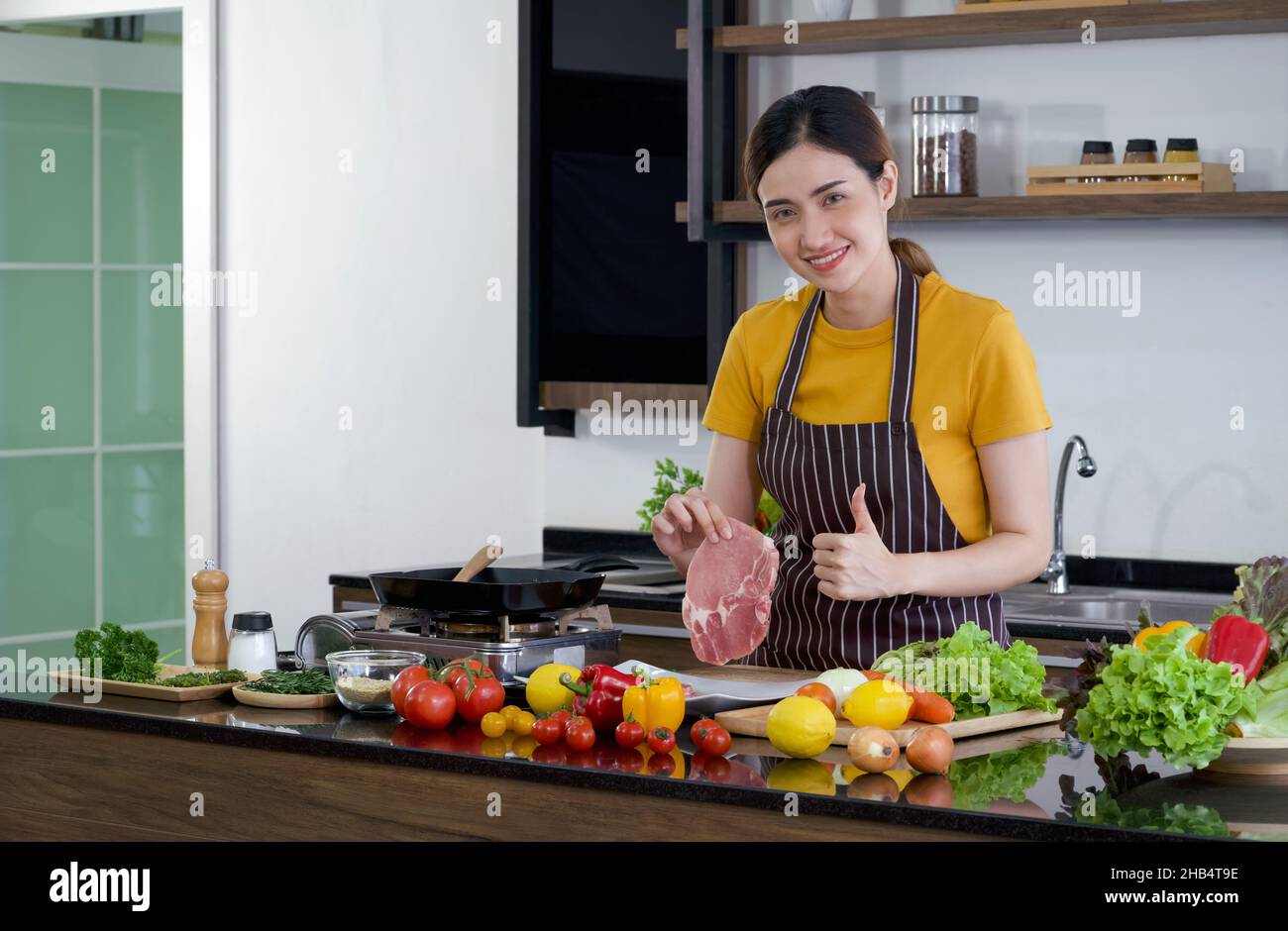 Young asian housewife dressed in an apron, smiled and raise finger thumb up while holding a piece of fresh steak. Morning atmosphere in a modern kitch Stock Photo