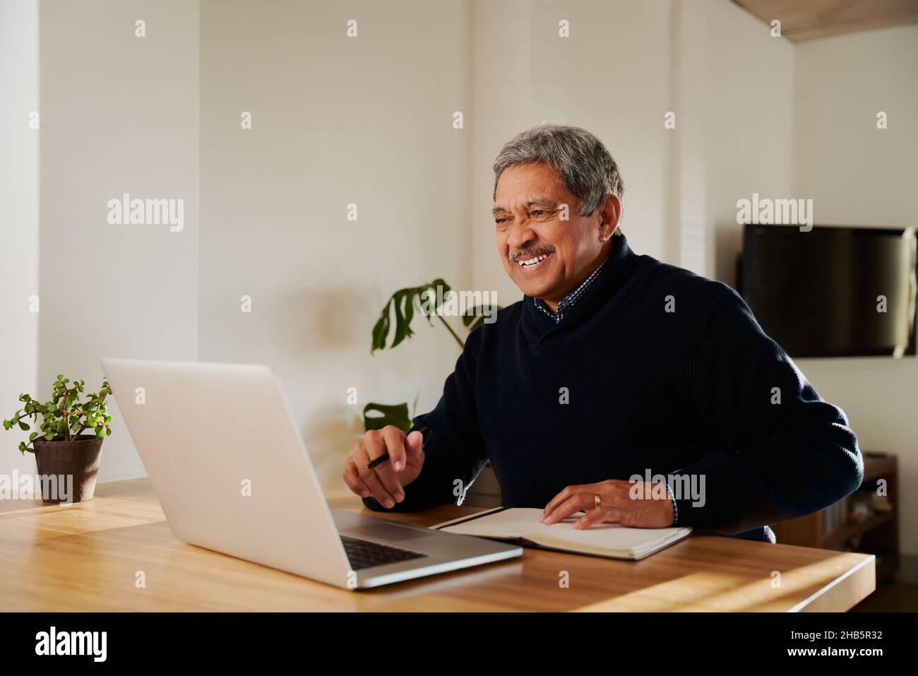 Multi-cultural elderly male smiling while on online call with family. Isolating in modern home, sitting at kitchen counter. Stock Photo