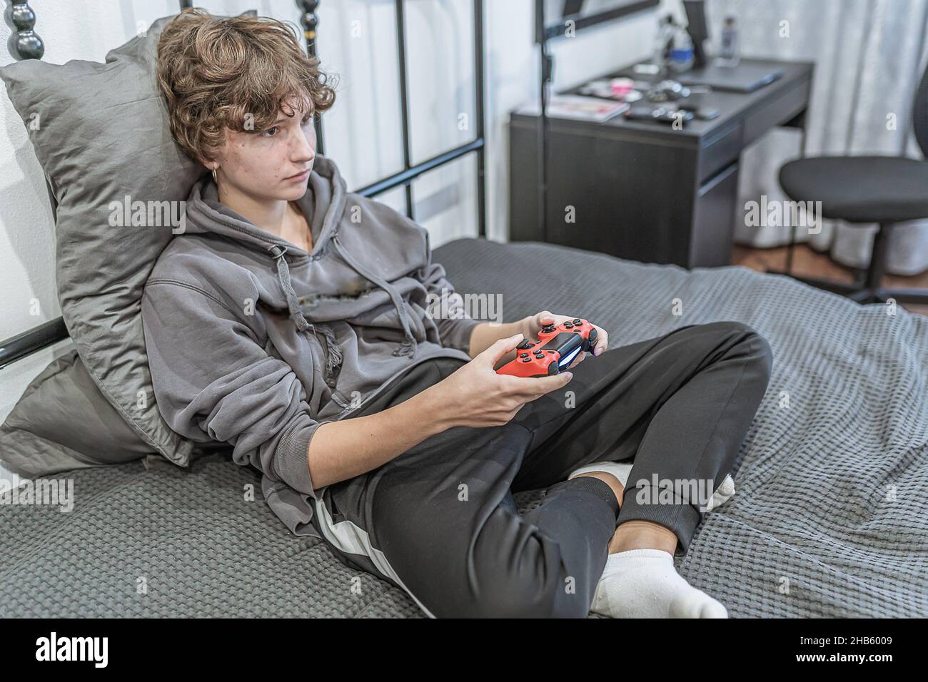 teenage girl with short brown curly hair, dressed in unkempt home clothes, sits on an unmade bed and plays on computer console. Passion for computer Stock Photo