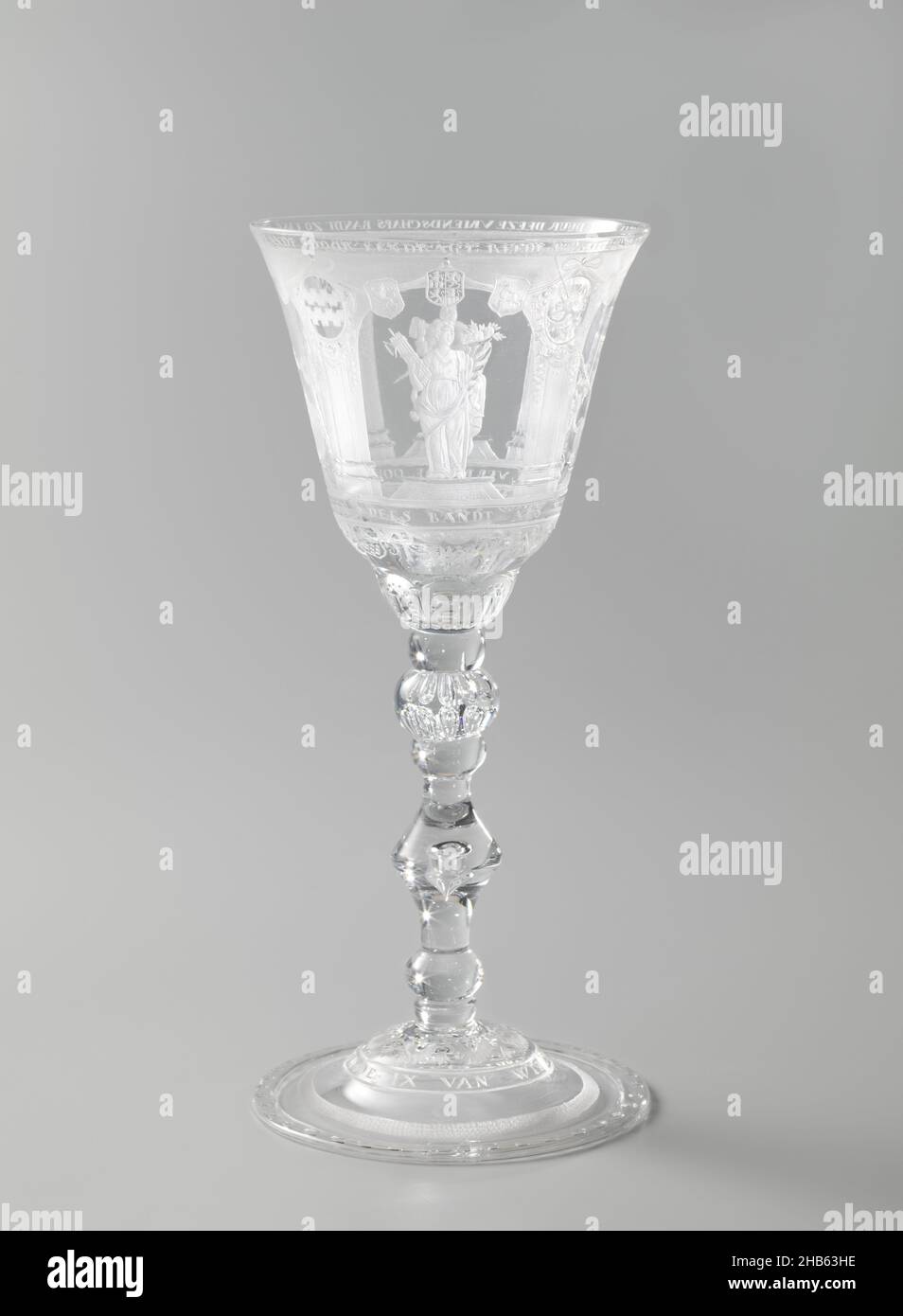 Wine glass, Hens cup of the Society The Ninths, Glass goblet decorated in carving. The base is trumpet-shaped, strongly hollowed in the center. The stem is composed of four smooth nodi between which is a lacy piece with one bubble and a thick nodus with two rows of bubbles. The chalice is conical with two curves at the bottom. The carving of the chalice: row of four arches with a figure standing on a pedestal. Man with fasces in left and horn of plenty in right hand. Crowned figure with sword in left hand and reins of lion behind him in right hand. Bearded man with heart in left hand and book Stock Photo