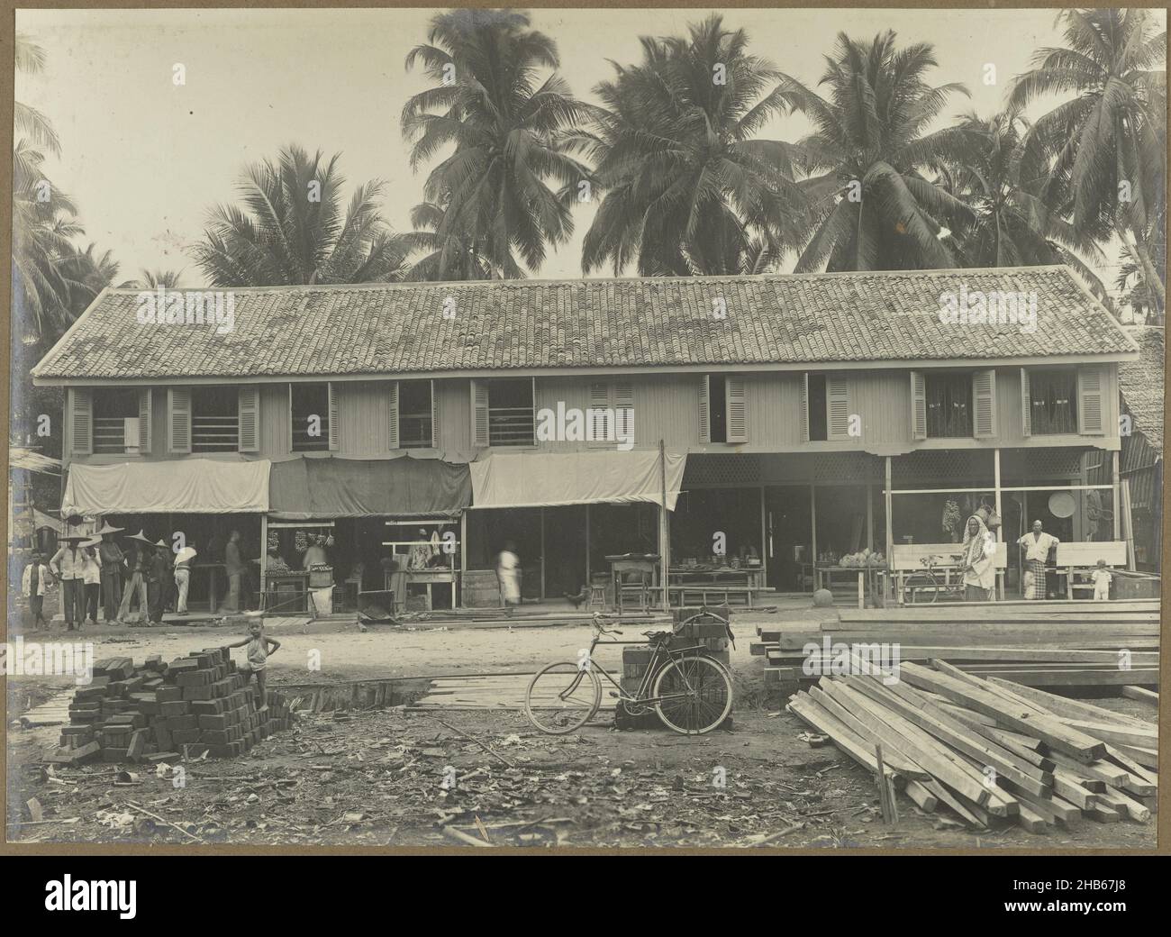 Commissioning of a store building, The commissioning of the finished block with several stores, probably in Medan. In the foreground a bicycle and piles of remaining building materials. Photo in the photo album of the Dutch architectural and contracting firm Bennink and Riphagen in Medan in the years ca. 1914-1919., anonymous, Medan, 1914 - 1919, photographic support, gelatin silver print, height 141 mm × width 197 mm Stock Photo