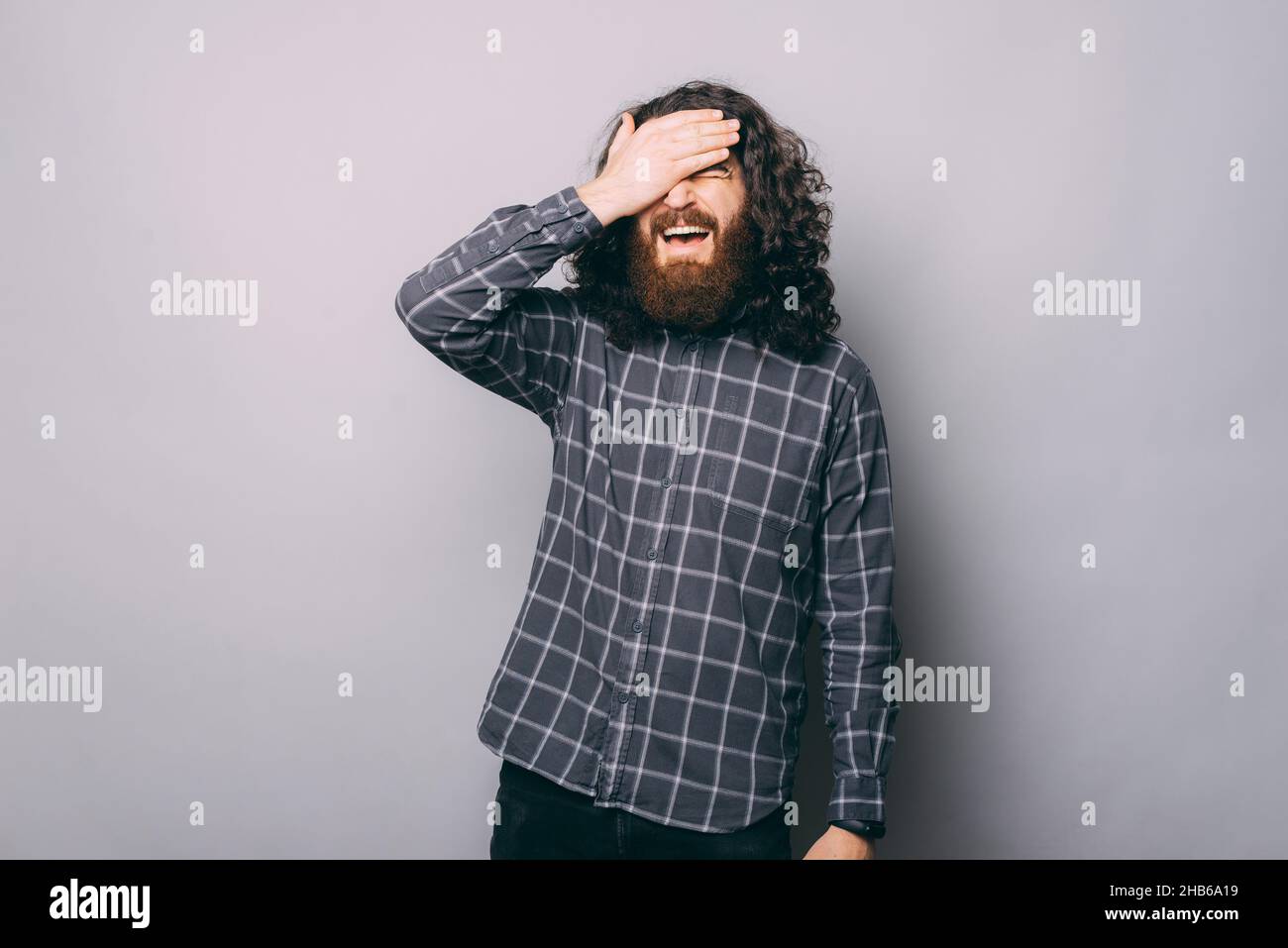 Confused bearded man in checkered shirt with closed eyes standing on gray background with hand on forehead in light studio Stock Photo