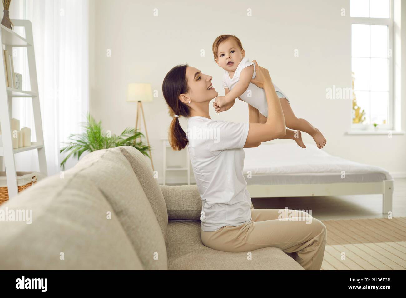Portrait of happy cheerful young woman holding her baby and playing with her at home. Stock Photo