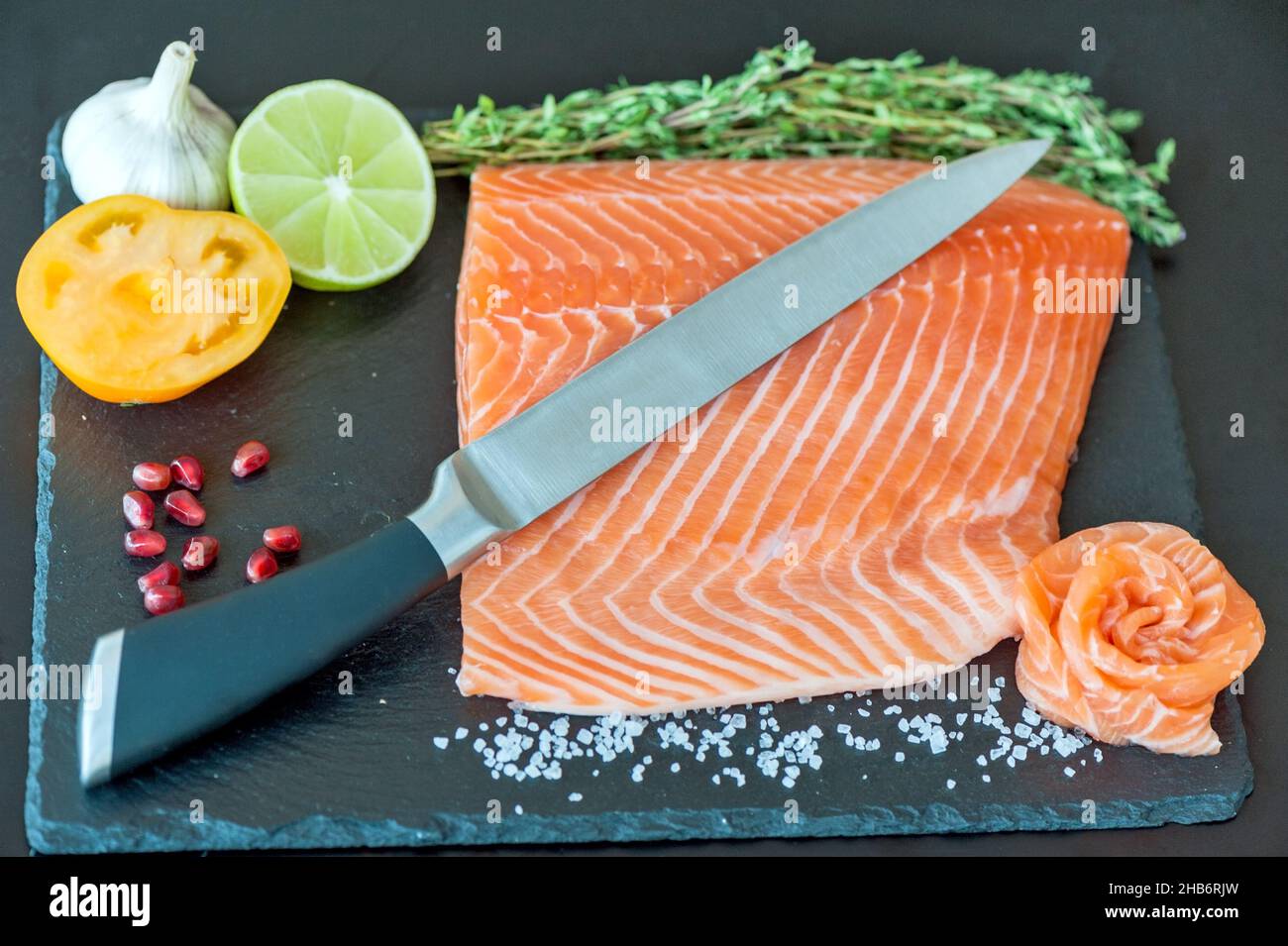 Hand chef using knife slice raw salmon on chopping block. Salmon fillet steak and seasonings on black cutting board. Stock Photo