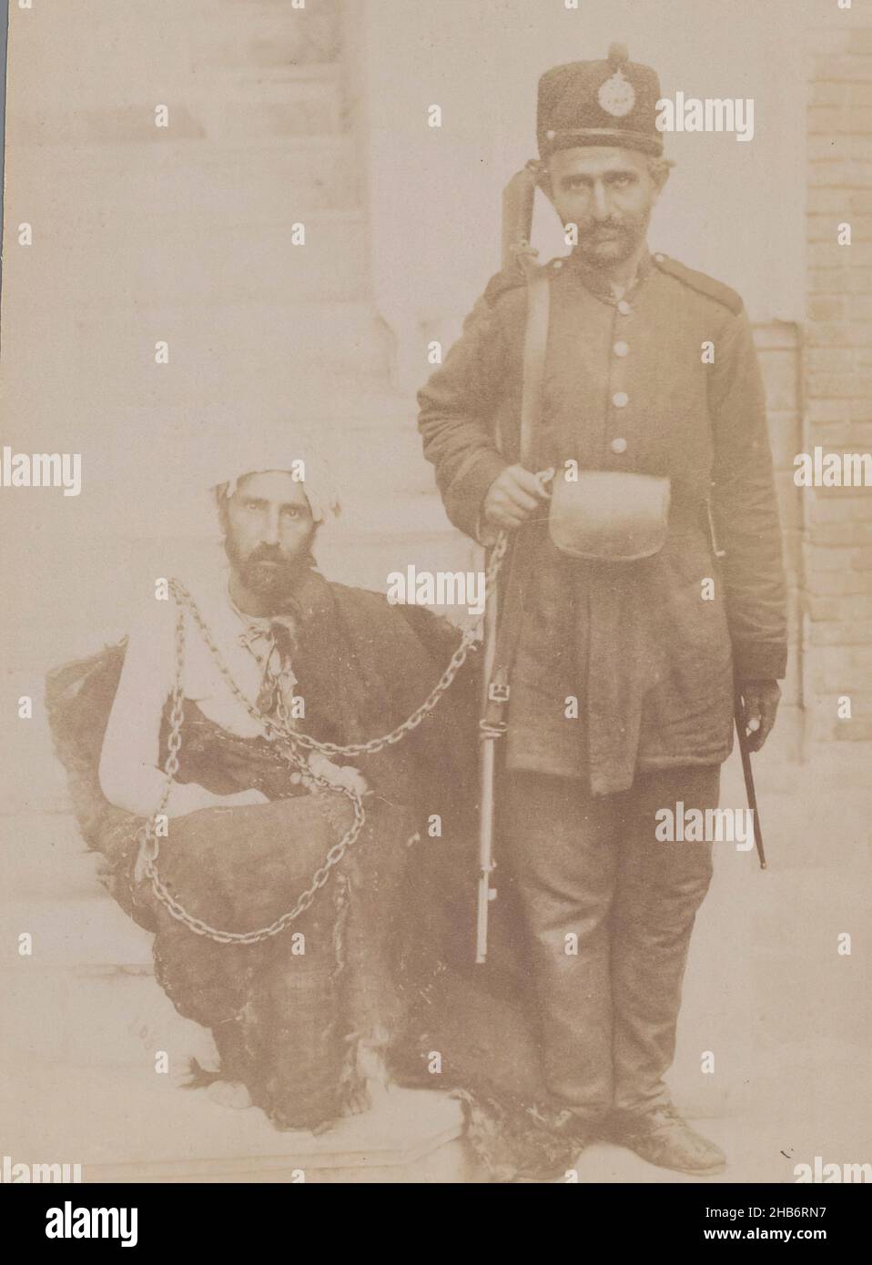 Portrait of two men: a prisoner and a guard, Iran, Frontal portrait of two men: a prisoner and a guard. The prisoner is chained with a long chain and crouches on the ground staring blankly and wrapped in a blanket. The guard in uniform stands and holds the chain in his hand, a rifle around the shoulder., Antoine Sevruguin, (attributed to), Iran, c. 1885 - c. 1910, paper, albumen print, height 204 mm, width 145 mm Stock Photo