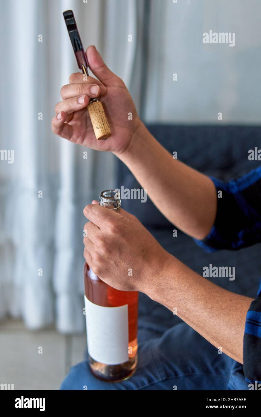 hands of latin man uncorking a bottle of rose wine while sitting on a sofa at home. Vertical Stock Photo