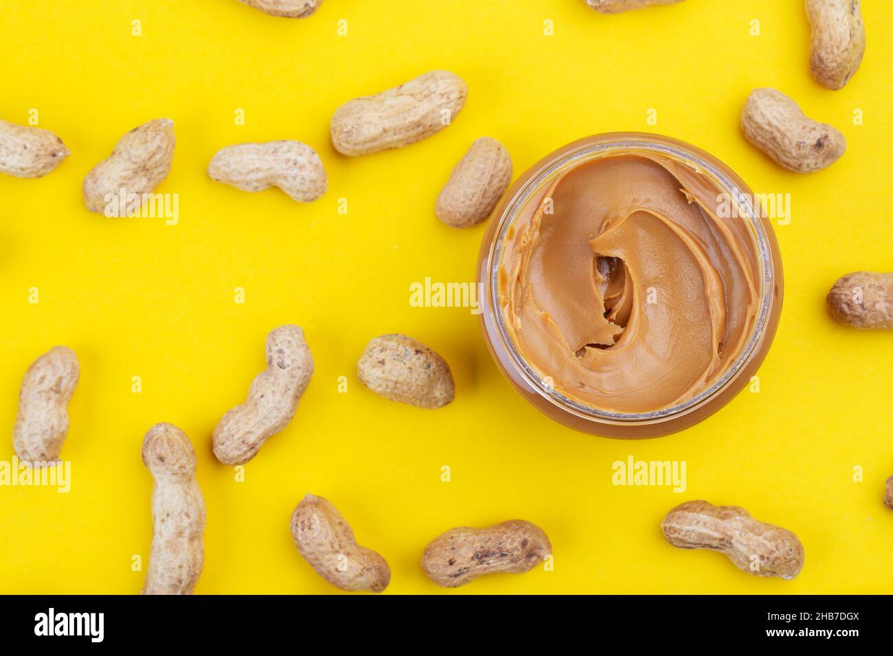 Peanut butter in a glass jar on a yellow background. Delicious and healthy breakfast. Arachis in a shell is scattered around the jar. Stock Photo