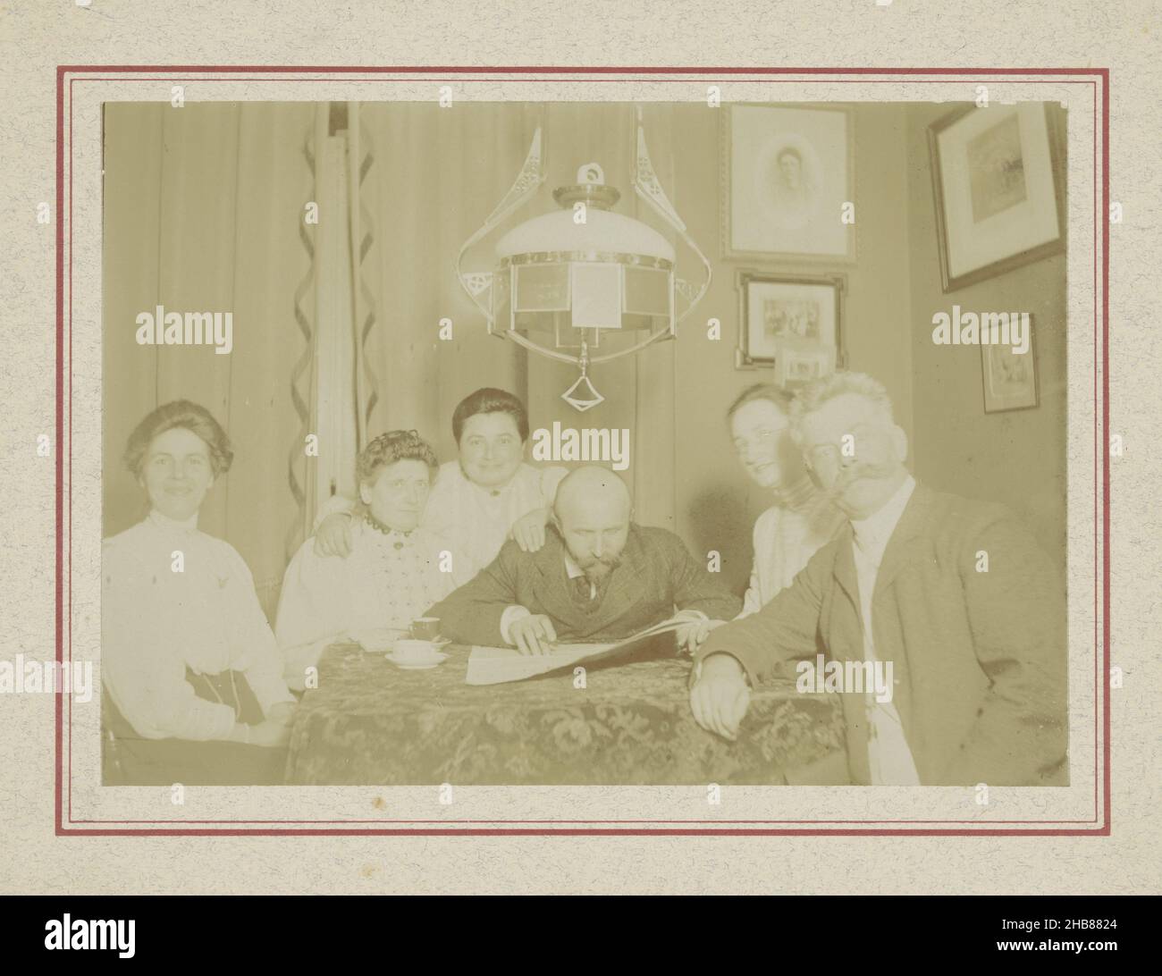 Group portrait around table in living room with photos on wall, Art-Nouveau lamp and man reading newspaper, anonymous, Netherlands, c. 1900 - c. 1910 Stock Photo