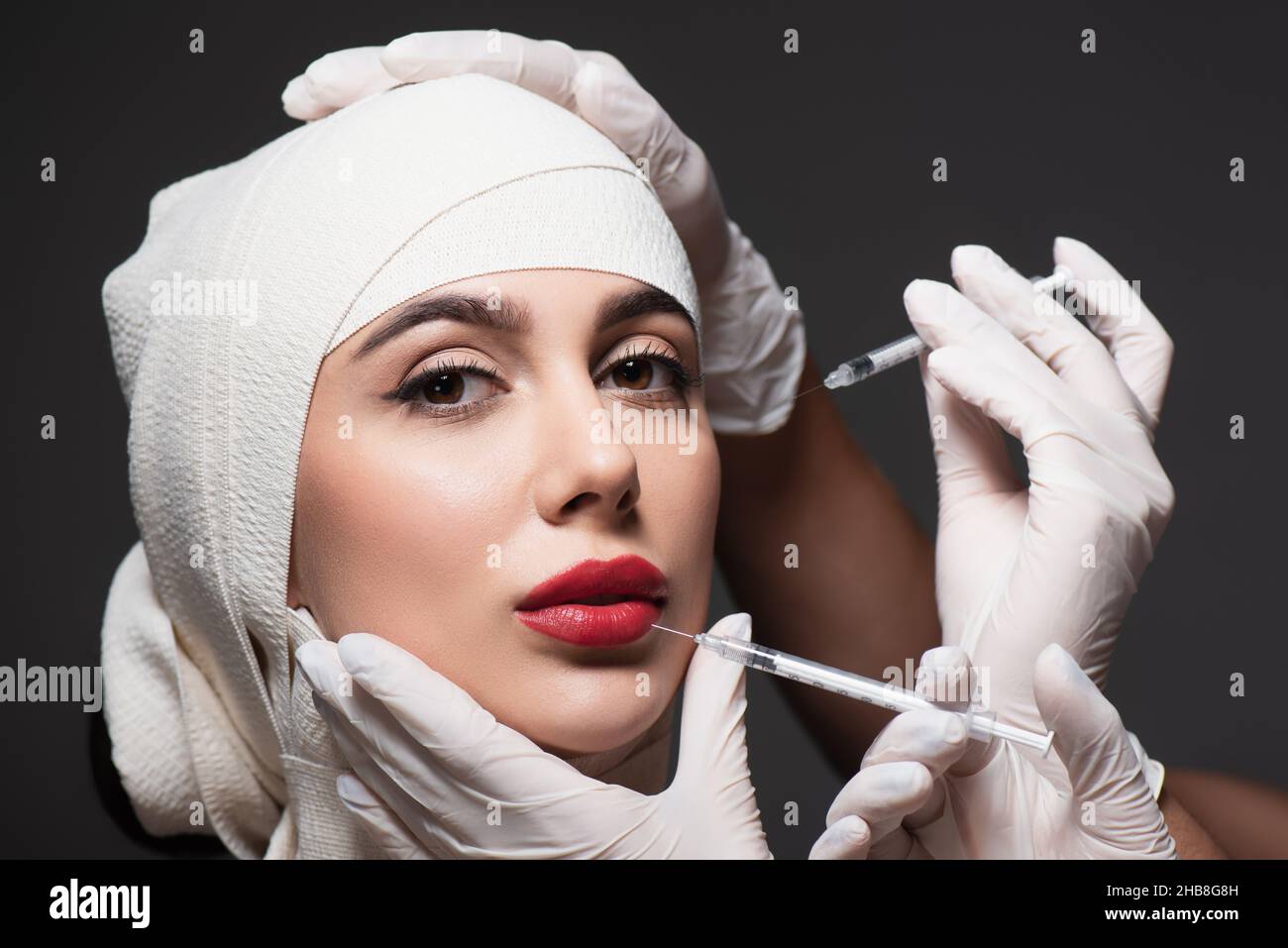 plastic surgeons holding syringes near patient in elastic bandage on head isolated on dark grey