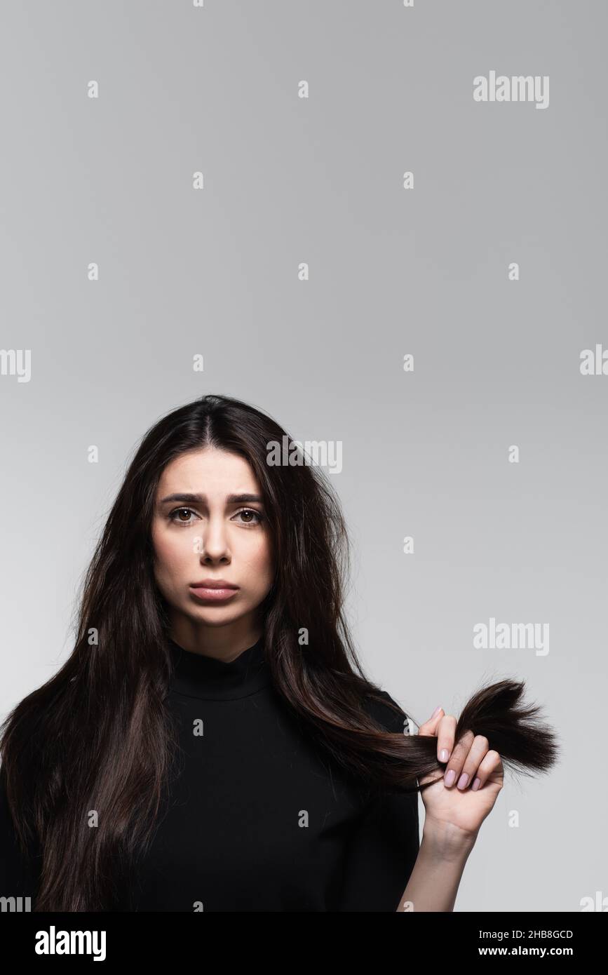 sad young woman in black turtleneck holding damaged hair isolated on grey Stock Photo