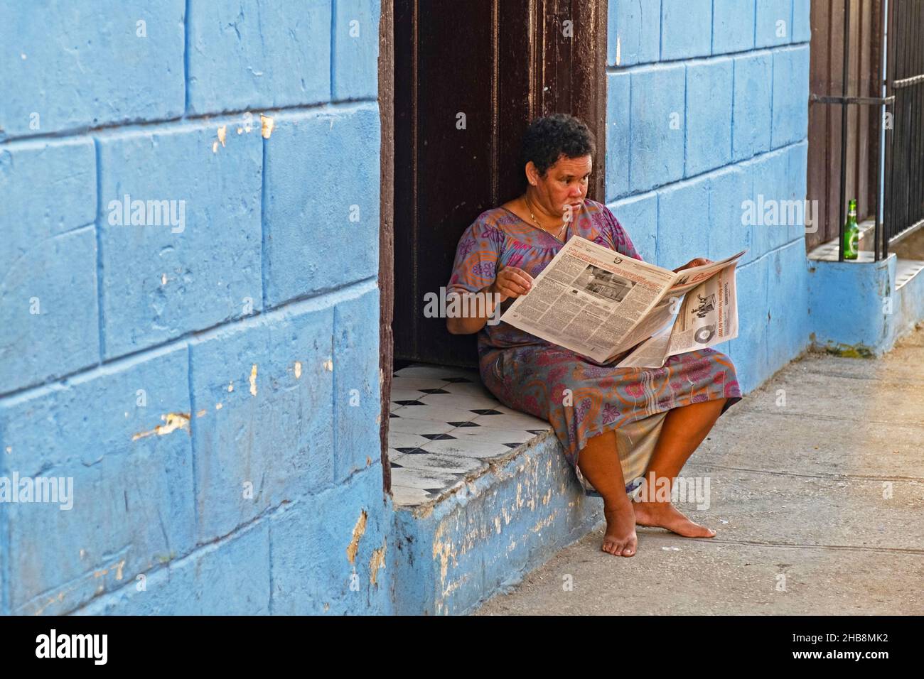 Cuban woman reading the Trabajadores, weekly Cuban trade union newspaper on the doorstep of her house in the city Sancti Spíritus on the island Cuba Stock Photo