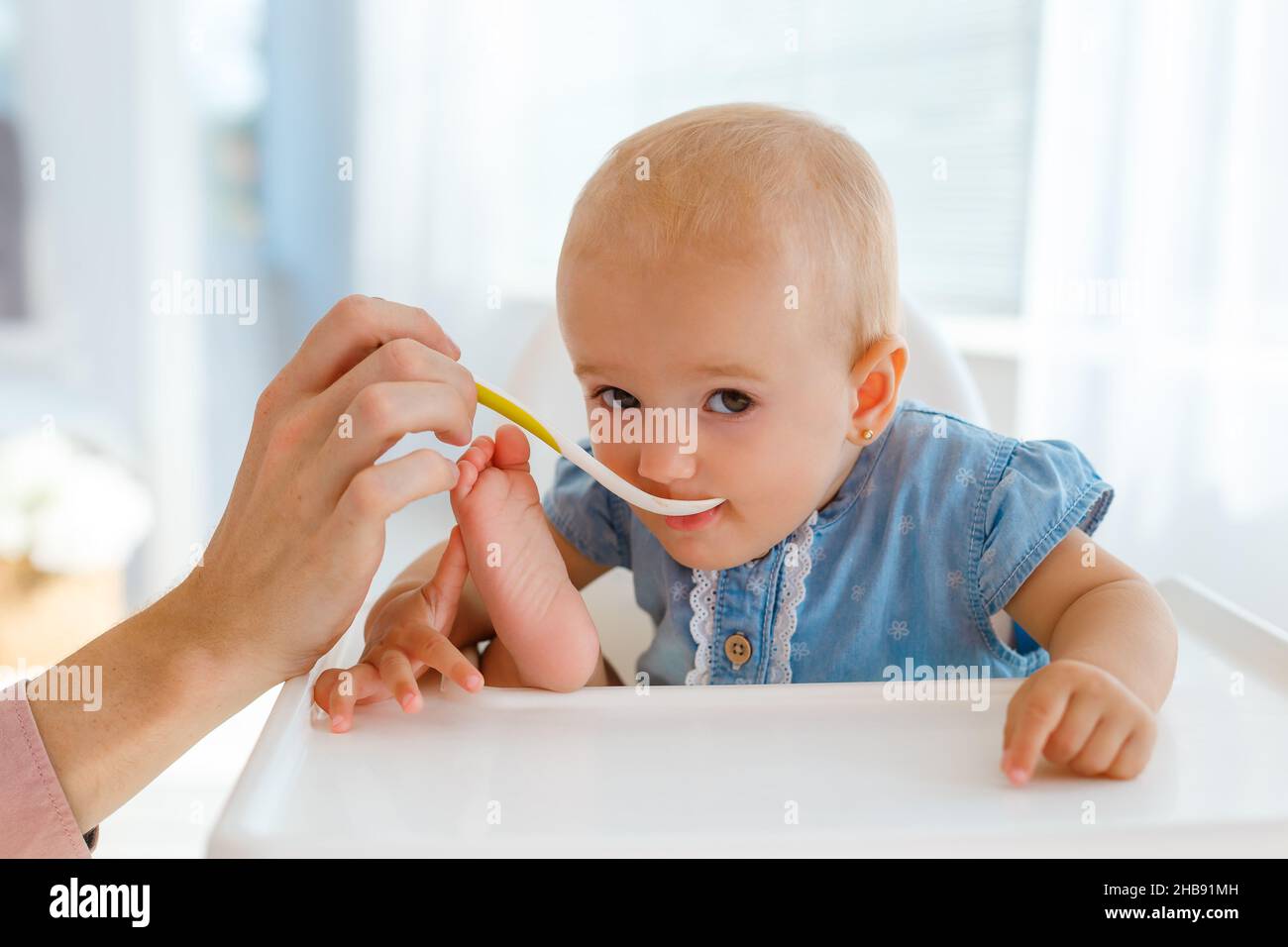 Very cute and funny baby with the spoon in hands eating kids nutrition - fruit puree. Smiling little girl eating in fun pose on white feeding table. C Stock Photo