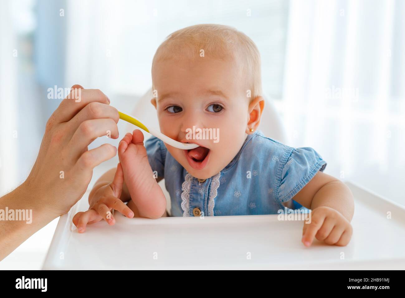 Very cute and funny baby with the spoon in hands eating kids nutrition - fruit puree. Smiling little girl eating in fun pose on white feeding table. C Stock Photo