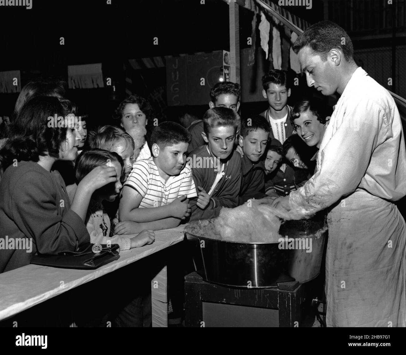 Young man in white shirt and apron making cotton candy while a bunch of children look on anxiously waiting for theirs. Circa 1950 Stock Photo