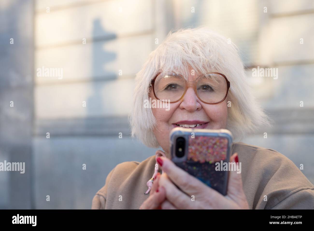 beautiful older woman with white hair looking at camera with smile and cell phone in her hands outdoors Stock Photo