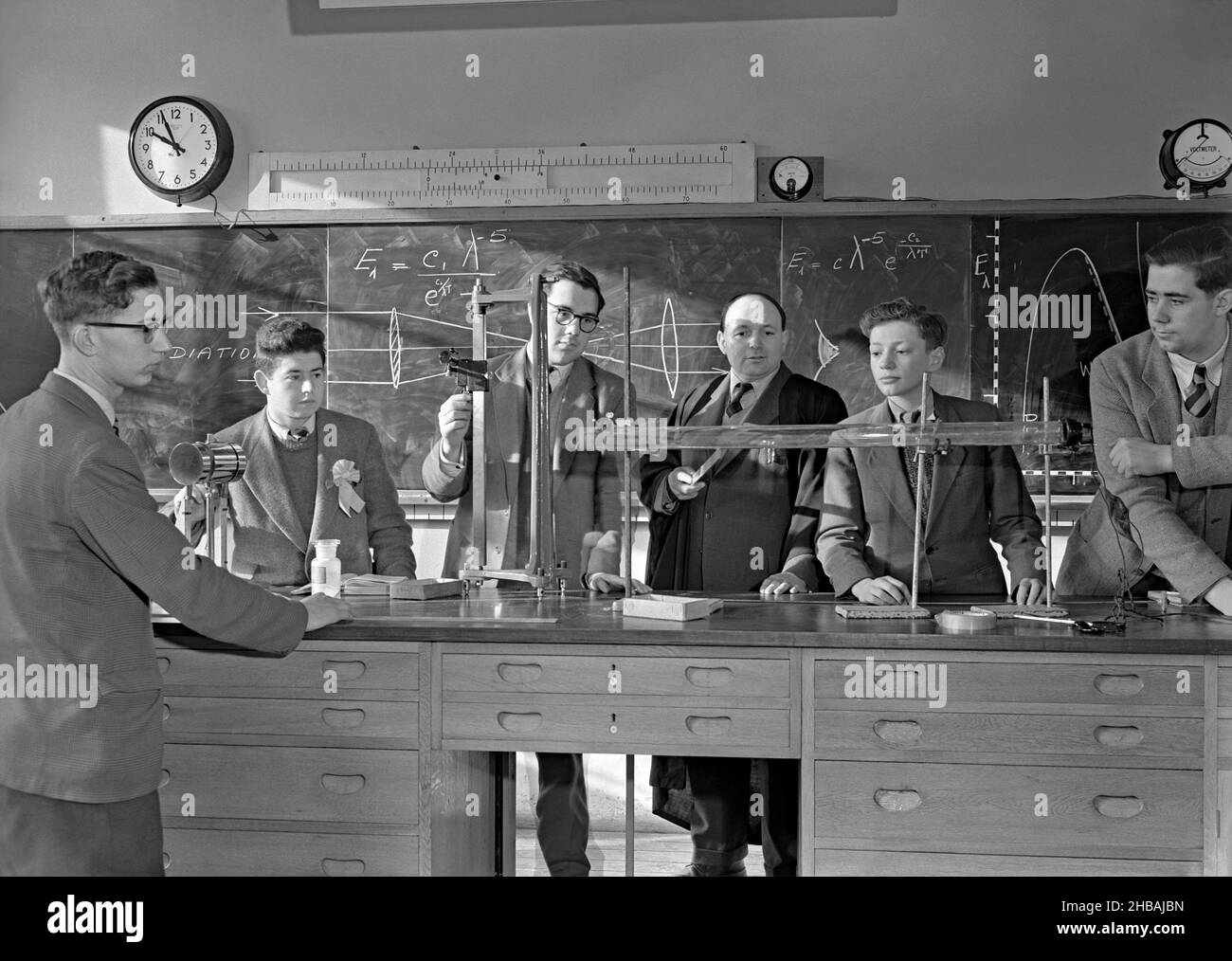 An experiment in the school science laboratory in Britain c.1960. The long tube the teacher and pupils are looking at is wired up – presumably this experiment is about current/light passing through a gaseous environment (the equations and drawings on the blackboard appear to be on radiation and the behaviour of light rays). Note the lack of protective eyewear at that time. This is taken from an old black and white negative – a vintage 1950s/60s photograph. Stock Photo