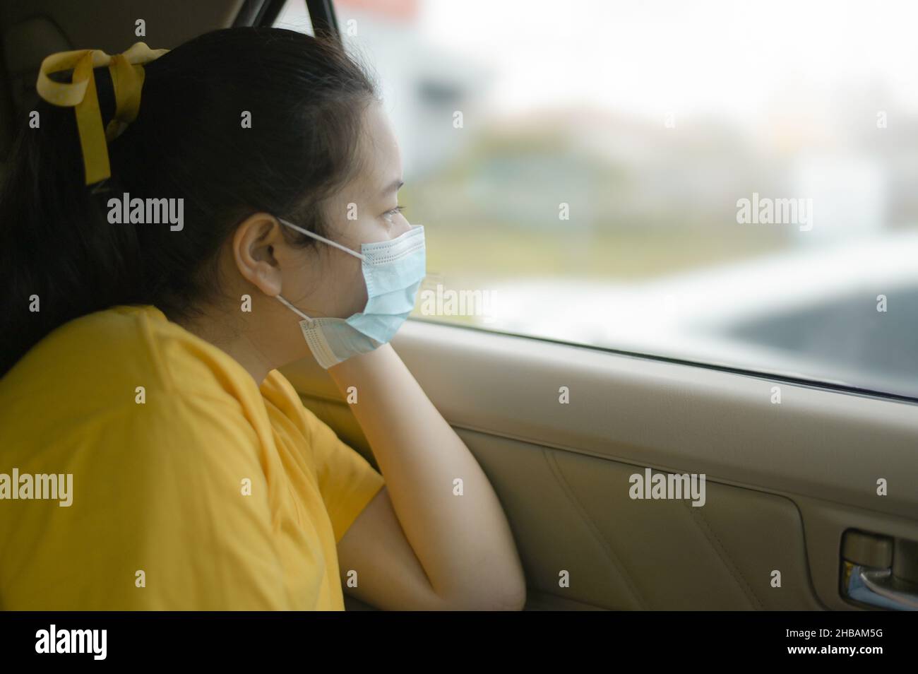A teenage girl in a mask sits and looks out of the car, Wearing yellow t-shirt sad eyes, virus outbreak. Stock Photo