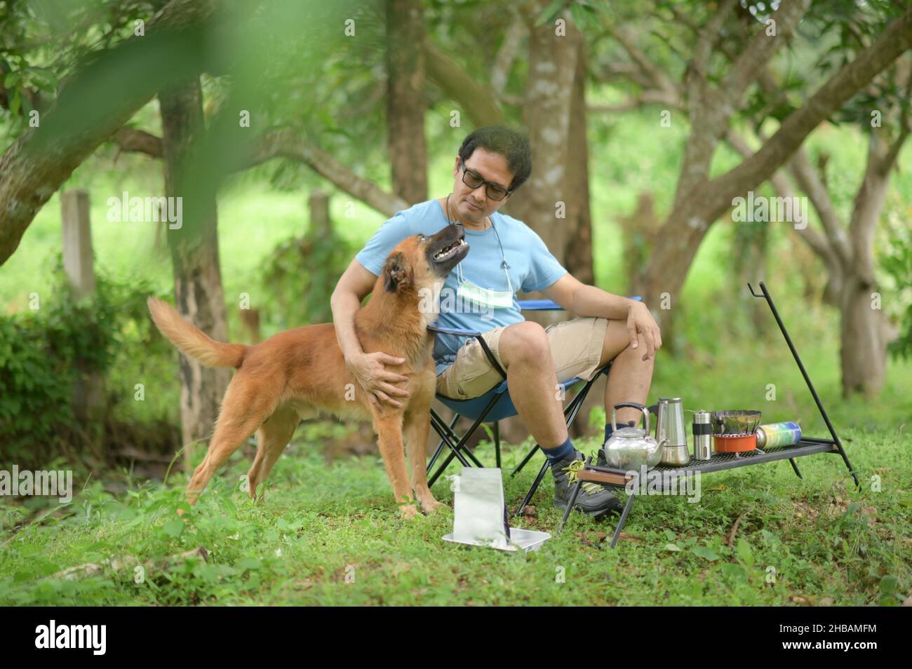 A man sits with his dog during camping, sits on a camping chair wearing glasses and a blue T-shirt, hugs the dog and makes a teasing face, coffee-maki Stock Photo