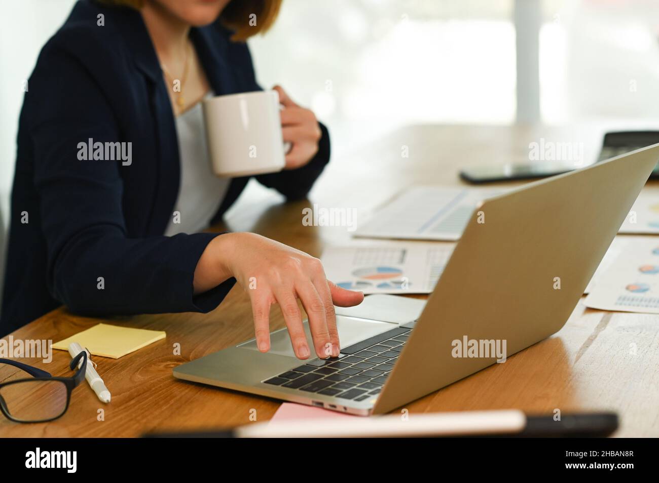 Woman in suit presses, hand on the laptop keyboard and holds coffee with the other hand, a young woman's hand is using laptop keyboard, drinking coffe Stock Photo