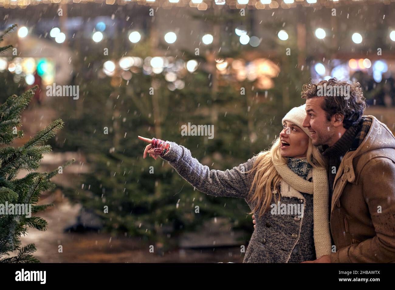 A young couple in love is commenting christmas trees while enjoying christmas holidays and walking the city on a beautiful snowy night. Christmas, New Stock Photo