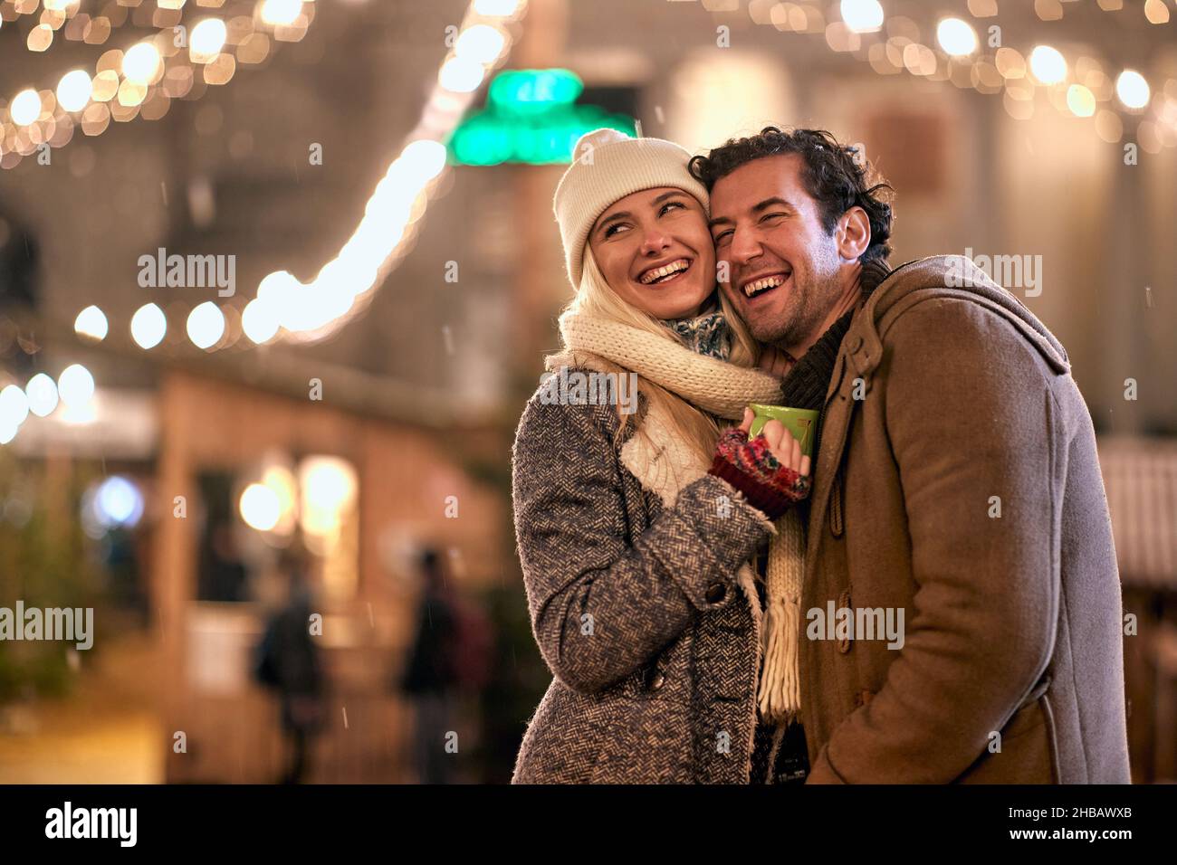 A young cheerful couple in a hug is standing under christmas light on a beautiful night in the city. Christmas, New Year, holiday, love Stock Photo