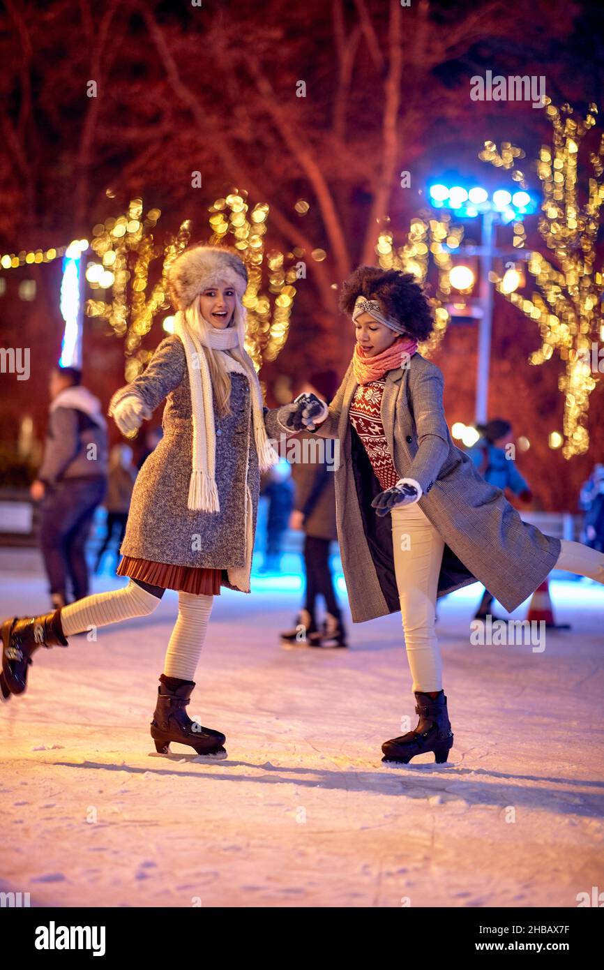 Two female friends are posing for a photo while skating at ice-skating rink during Christmas holidays on a beautiful night in the city. Christmas, New Stock Photo
