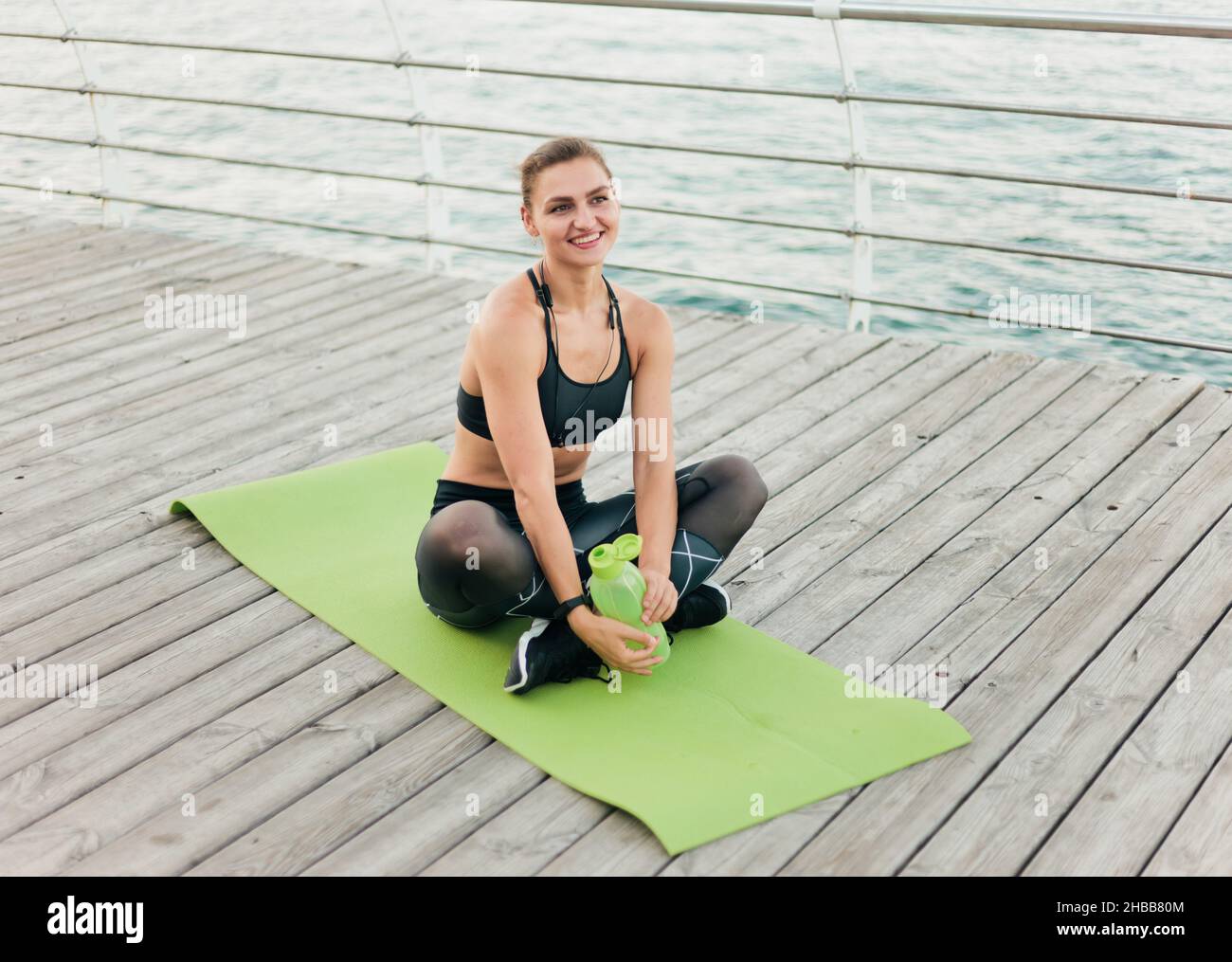 Young happy sport woman in sportwear sitting on mat at wooden terrace ...