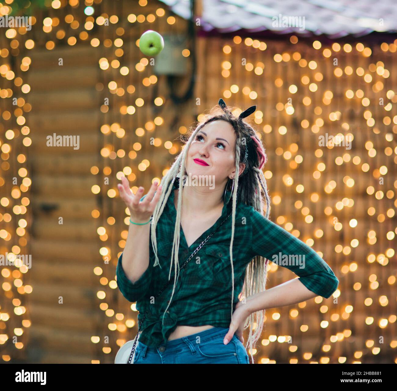 Inordinate young woman with dreadlocks hairstyle and fashionable summer clothes throws an apple against the background of garlands Stock Photo