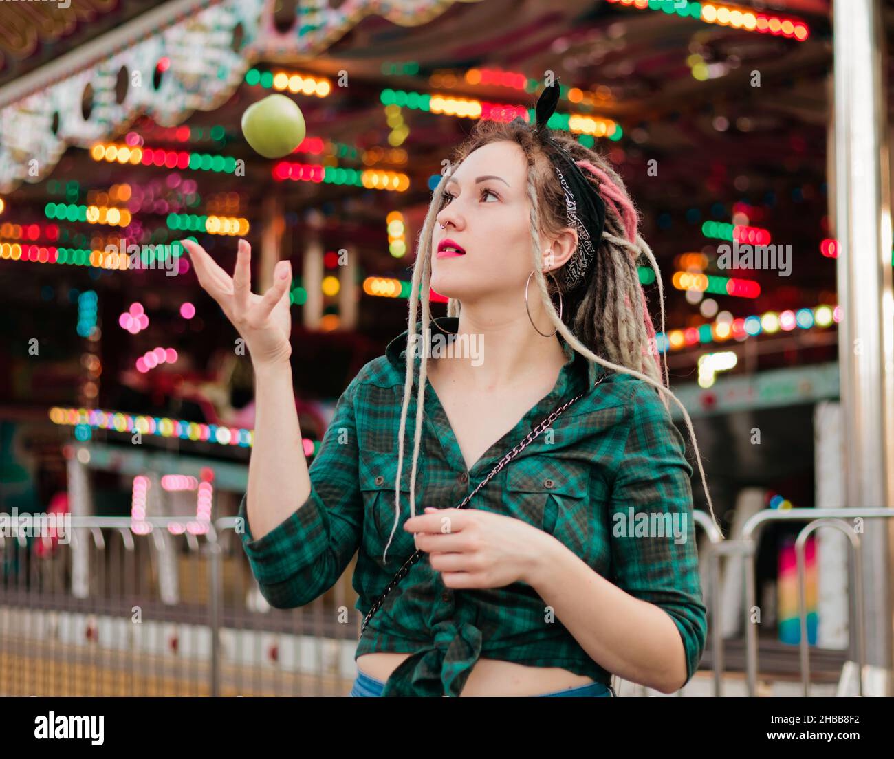 Inordinate young woman with dreadlocks hairstyle and fashionable summer clothes throws apple in amusement park. Stock Photo