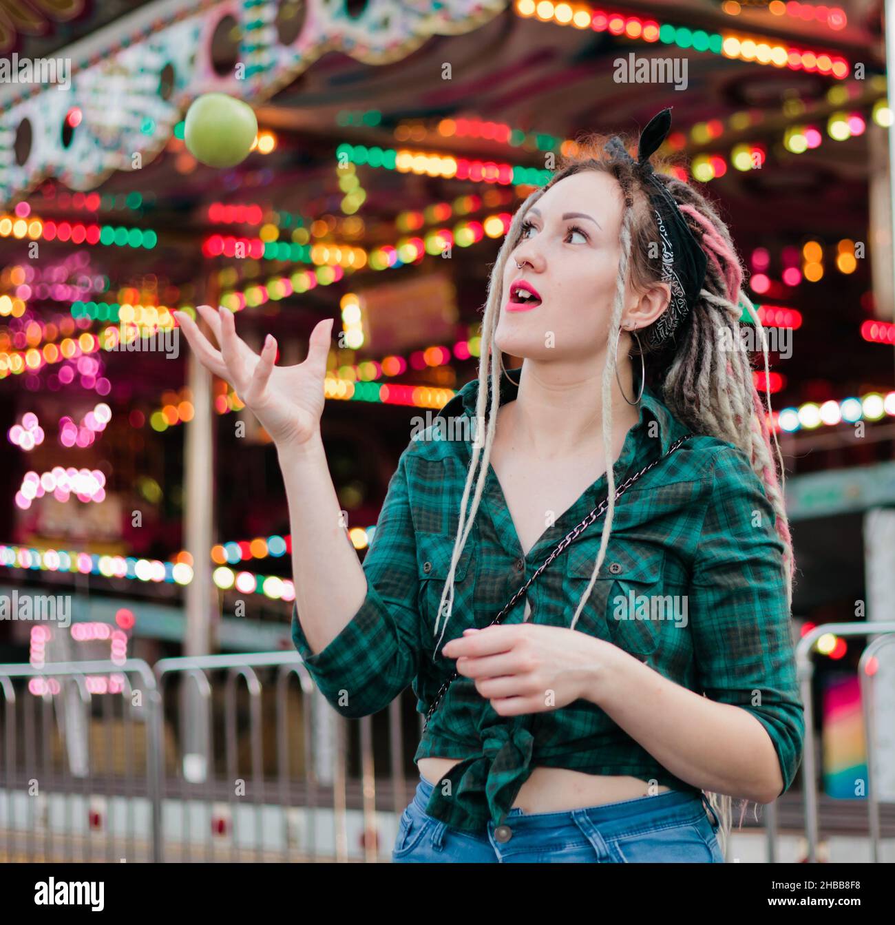 Inordinate young woman with dreadlocks hairstyle and fashionable summer clothes throws apple in amusement park. Stock Photo