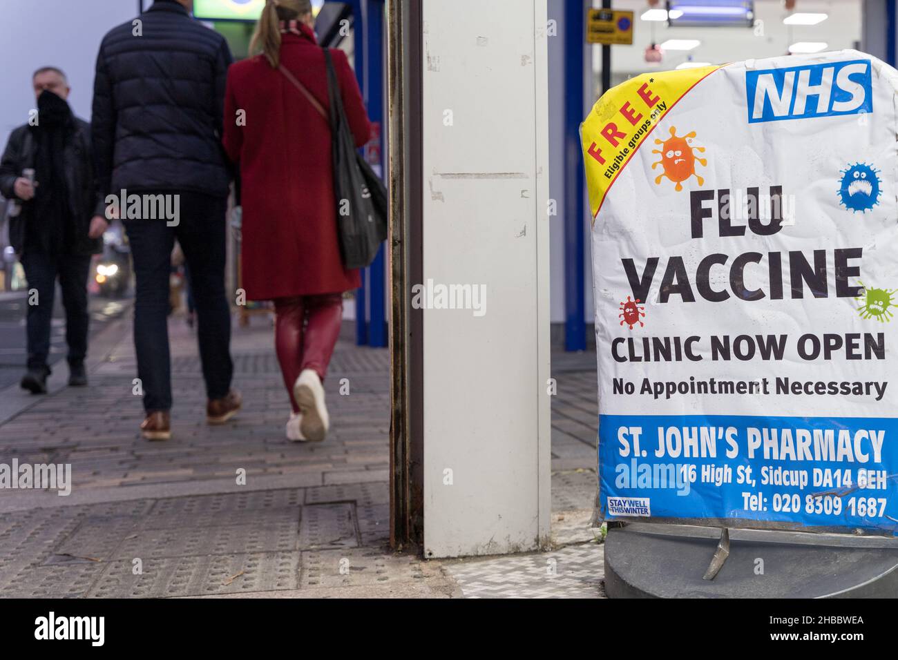 Christmas shoppers walk past Flu Vaccine poster offering free flu jab to those eligible people at Sidcup pharmacy London England Stock Photo