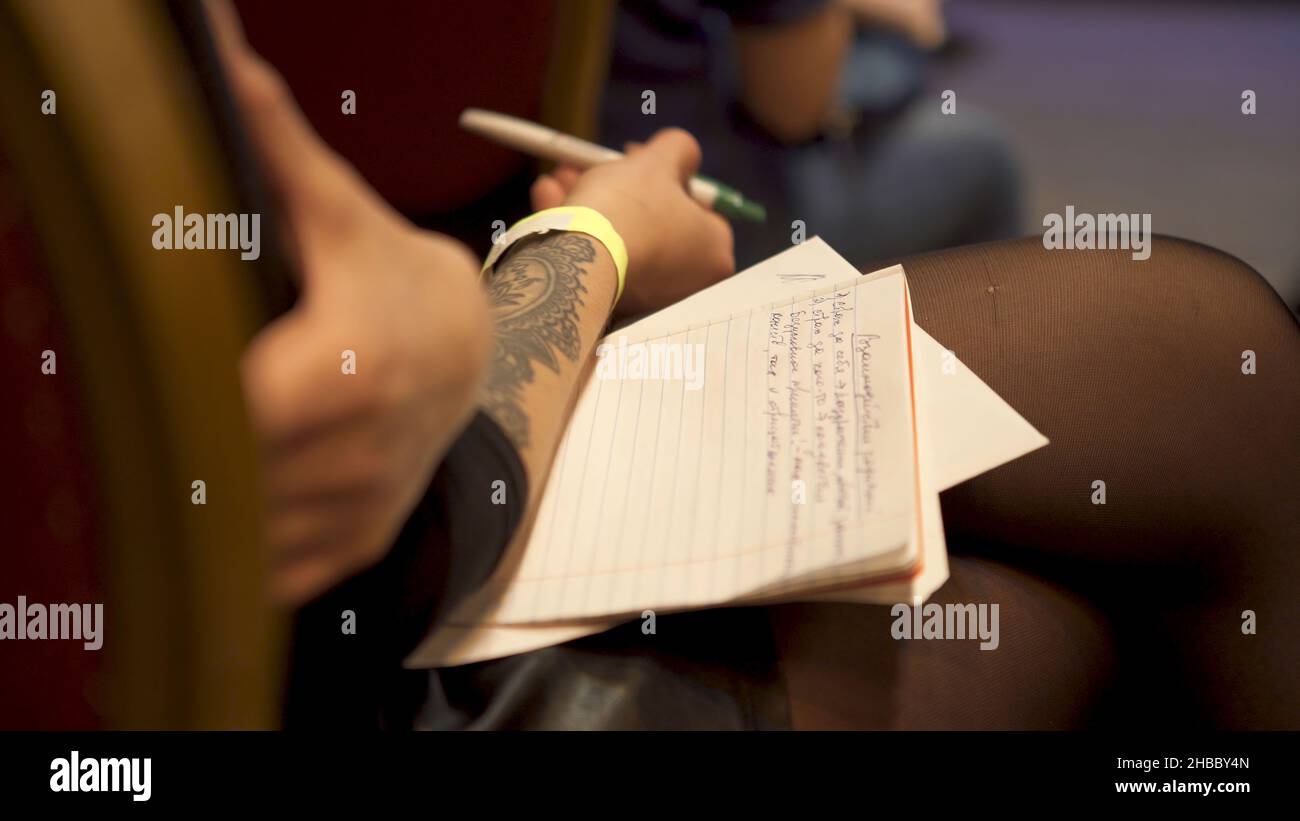 Close up for woman sitting in a chair with a copybook on her knees and a pen in her hand. Young girl with a tattoo on her hand holding a pen and noteb Stock Photo