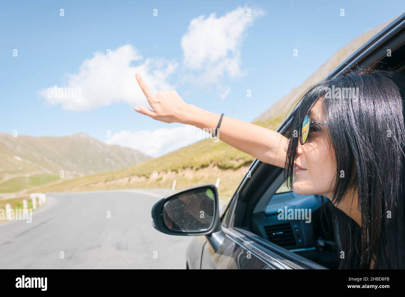 Female caucasian woman from car window points finger up to left with asphalt road background. Copypaste raod trip concept. Stock Photo