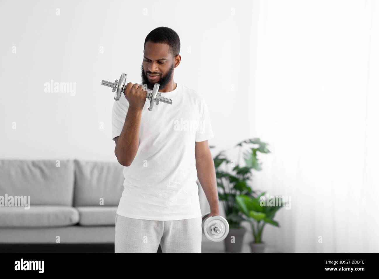 Serious young black sportsman man in white clothes doing exercise with dumbbells in minimalist living room Stock Photo